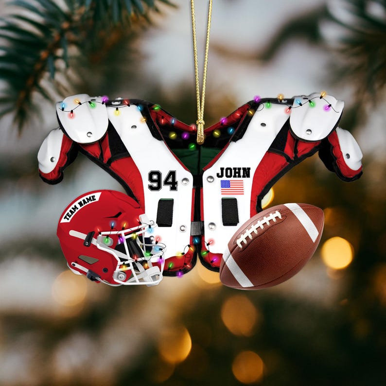 K&ouml;nnte beinhalten: Ein weihnachtlicher Football-Ornament. Es zeigt ein rot-wei&szlig;es Schulterpolster-Design, einen roten Helm mit dem Text "TEAM NAME" und einen braunen Football. Das Ornament enth&auml;lt die Nummer "94", den Namen "JOHN" und eine amerikanische Flagge.