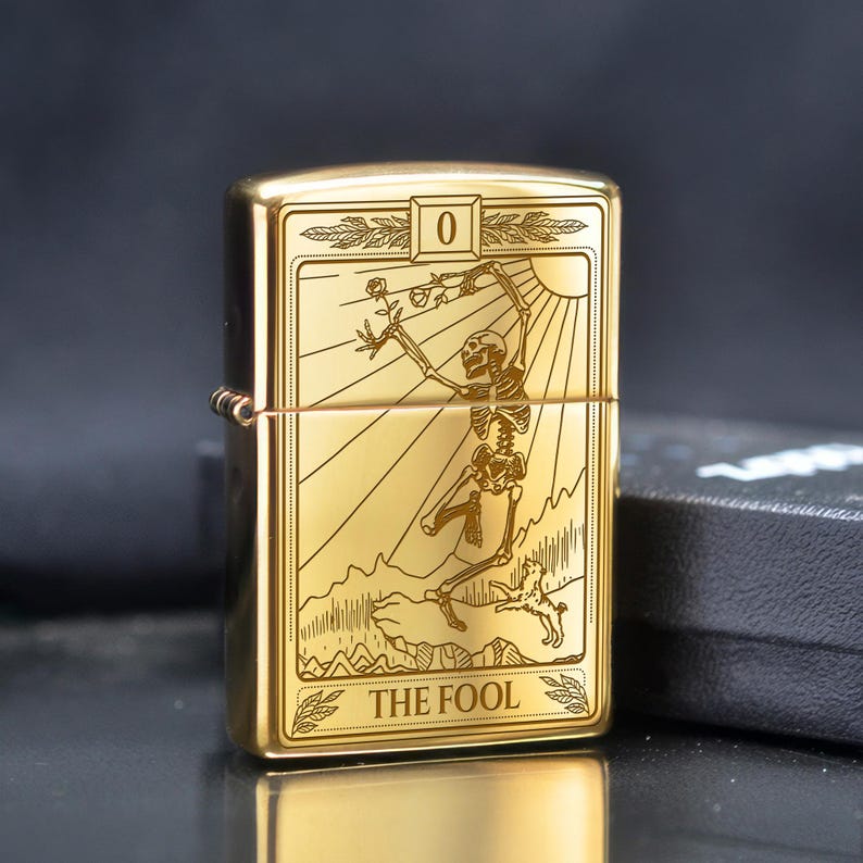 May include: Gold-colored lighter with a tarot card design. The card features a skeleton figure holding a rose, with the text "THE FOOL" at the bottom. The lighter has a rectangular shape and a hinged lid.