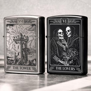 May include: Two metal lighters, one silver and one black, each featuring tarot card designs. The silver lighter displays "The Tower" card with a tower illustration. The black lighter shows "The Lovers" card with skeleton figures. Both lighters have a rectangular shape.
