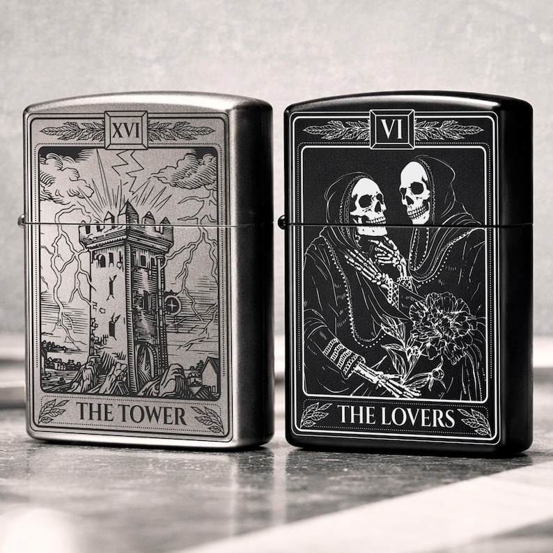 May include: Two metal lighters, one silver and one black, each featuring tarot card designs. The silver lighter displays "The Tower" card with a tower illustration. The black lighter shows "The Lovers" card with skeleton figures. Both lighters have a rectangular shape.