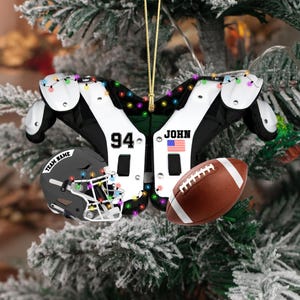 K&ouml;nnte beinhalten: Ein weihnachtlicher Christbaumschmuck mit Football-Motiv. Er zeigt einen Helm, Schulterpolster und einen Football. Die Schulterpolster sind wei&szlig; und schwarz mit der Nummer 94 und dem Namen "JOHN" mit einer amerikanischen Flagge. Der Helm hat den Text "TEAM NAME".