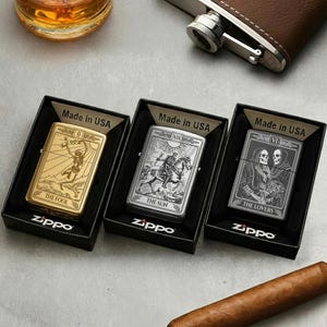 May include: Three Zippo lighters in black boxes, each featuring a different tarot card design: "The Fool" in gold, "The Sun" in silver, and "The Lovers" in gray. The lighters are marked "Made in USA" and have the Zippo logo.