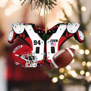 K&ouml;nnte beinhalten: Fu&szlig;ball-Weihnachtsschmuck mit einem roten Helm mit dem Text "TEAM NAME", einem braunen Football und einem wei&szlig;-roten Schulterpolster-Design mit der Nummer "94", dem Namen "JOHN" und einer amerikanischen Flagge. Der Schmuck ist mit Lichterketten verziert.