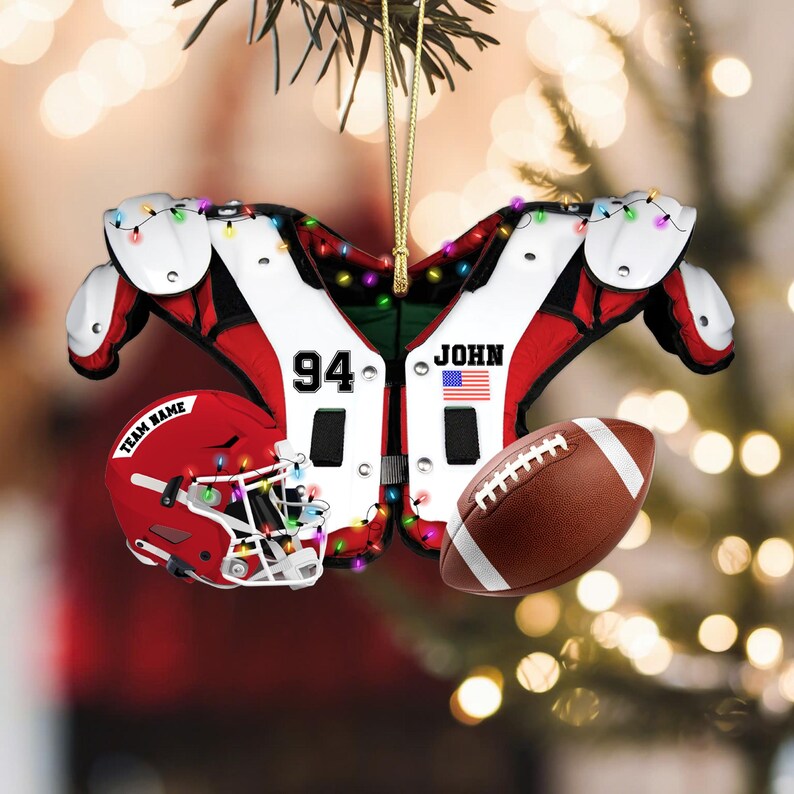 K&ouml;nnte beinhalten: Fu&szlig;ball-Weihnachtsschmuck mit einem roten Helm mit dem Text "TEAM NAME", einem braunen Football und einem wei&szlig;-roten Schulterpolster-Design mit der Nummer "94", dem Namen "JOHN" und einer amerikanischen Flagge. Der Schmuck ist mit Lichterketten verziert.