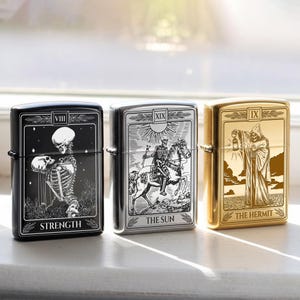 May include: Three metal lighters with tarot card designs. The black lighter features a skeleton and the word "STRENGTH". The silver lighter depicts a skeleton on horseback with "THE SUN". The gold lighter shows a cloaked figure with "THE HERMIT".
