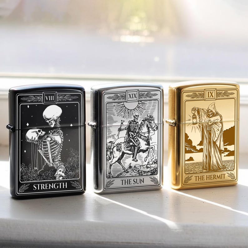 May include: Three metal lighters with tarot card designs. The black lighter features a skeleton and the word "STRENGTH". The silver lighter depicts a skeleton on horseback with "THE SUN". The gold lighter shows a cloaked figure with "THE HERMIT".