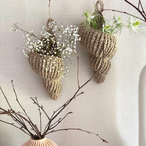Könnte beinhalten: Zwei hängende, gewebte, muschelförmige Pflanzgefäße, gefüllt mit weißen Blumen. Die Pflanzgefäße sind mit Schnur aufgehängt und bestehen aus einem natürlichen, hellbraunen Material. Der Hintergrund ist eine neutrale, cremefarbene Farbe, mit einer Vase und Ästen im Vordergrund.