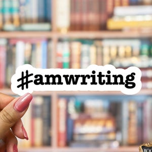 May include: A white die-cut sticker featuring the black text #amwriting in a bold, rounded serif font. The sticker has a thin white border around the lettering. In the bottom right corner, a small black rectangular logo reads The Bookish Swag Shop.