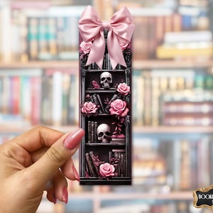 May include: A decorative bookmark featuring a gothic bookshelf design with skulls, roses, and books. The bookmark is adorned with a pink bow at the top. The Bookish Swag Shop logo is visible at the bottom.