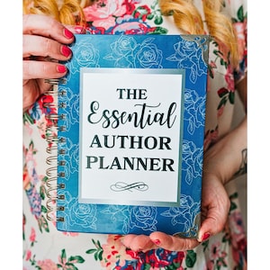 May include: A spiral-bound planner with a deep blue cover featuring a white rose line art pattern. The center white label displays the text THE Essential AUTHOR PLANNER in black lettering. The planner includes metallic corner protectors.