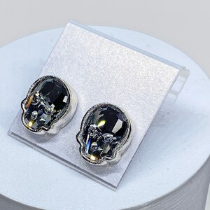 May include: A pair of silver skull stud earrings with black crystal accents.