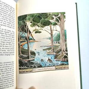 K&ouml;nnte beinhalten: Eine Illustration eines Hobbits, Bilbo Beutlin, der mit einem Boot einen Fluss hinunterf&auml;hrt. Der Fluss ist von B&auml;umen umgeben und der Himmel ist blassblau. Der Text "Bilbo kommt zu den H&uuml;tten der Ruhe-Elfen" befindet sich am unteren Rand des Bildes.