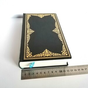 May include: A black hardcover book with a gold ornate design on the cover. The book is closed and lying flat on a white surface.