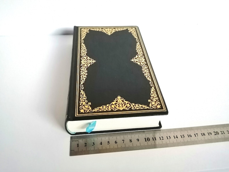 May include: A black hardcover book with a gold ornate design on the cover. The book is closed and lying flat on a white surface.