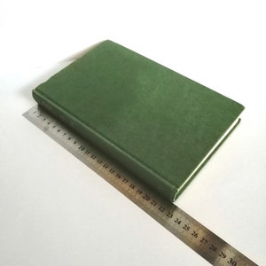 K&ouml;nnte beinhalten: Ein gr&uuml;nes Hardcover-Buch liegt auf einer wei&szlig;en Oberfl&auml;che mit einem Metalllineal daneben. Das Lineal ist mit Zahlen von 1 bis 33 gekennzeichnet.
