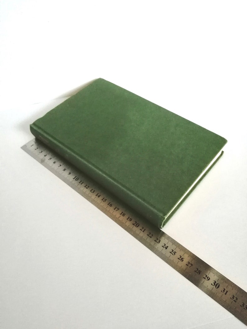 K&ouml;nnte beinhalten: Ein gr&uuml;nes Hardcover-Buch liegt auf einer wei&szlig;en Oberfl&auml;che mit einem Metalllineal daneben. Das Lineal ist mit Zahlen von 1 bis 33 gekennzeichnet.