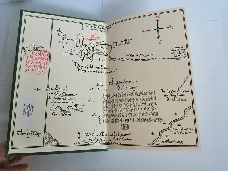 K&ouml;nnte beinhalten: Eine Schwarzwei&szlig;karte eines fiktiven Landes mit dem Text "Thror's Map" in der linken unteren Ecke. Die Karte zeigt eine Bergkette, einen Fluss und einen See. Der Text "Here of old was Thorin King under the Mountain" ist in roter Tinte geschrieben.