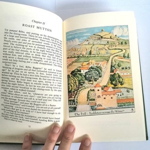 K&ouml;nnte beinhalten: Ein offenes Buch mit einer farbenfrohen Illustration eines gr&uuml;nen H&uuml;gels mit einem gewundenen Pfad, der zu einem kleinen Dorf f&uuml;hrt. Das Buch tr&auml;gt den Titel "The Hill: Hobbiton-across-the-Water". Der Text auf der Seite beschreibt eine Szene aus dem Buch "Der Hobbit".