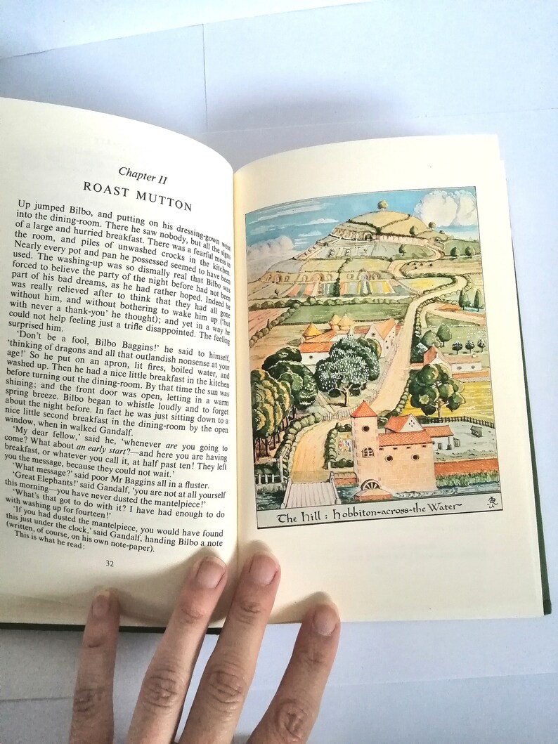 K&ouml;nnte beinhalten: Ein offenes Buch mit einer farbenfrohen Illustration eines gr&uuml;nen H&uuml;gels mit einem gewundenen Pfad, der zu einem kleinen Dorf f&uuml;hrt. Das Buch tr&auml;gt den Titel "The Hill: Hobbiton-across-the-Water". Der Text auf der Seite beschreibt eine Szene aus dem Buch "Der Hobbit".