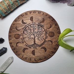 May include: A wooden circular divination board with a tree of life design and moon phases around the edge. The board has letters of the alphabet, numbers, and the words "Yes", "Maybe", "No", "Wait", "Rephrase", and "Try Again".