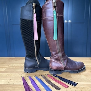 May include: A black tall boot with a textured panel and a pink tassel on the zip, and a brown tall boot with a green tassel on the zip. Six additional leather tassels in purple, blue, brown, red, and black are arranged in a row below.