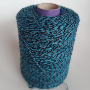 Peut inclure: Une grosse bobine de fil aux tons de bleu sarcelle, noir et gris. Le fil est étroitement enroulé autour d'un noyau en carton avec un bord violet. Un brin de fil s'étend du bas de la bobine.