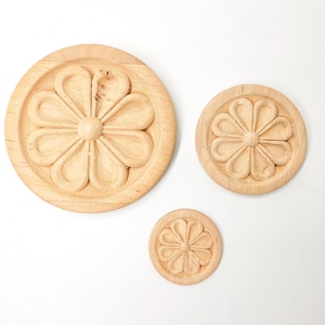 May include: Three wooden round ornaments with a carved flower design. The ornaments are different sizes and are arranged in a row on a white background.