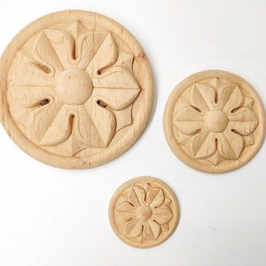 May include: Three wooden round ornaments with a carved floral design. The ornaments are different sizes and are arranged in a row on a white surface.