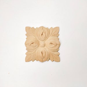 Puede incluir: Un adorno cuadrado de madera tallada con un dise&ntilde;o floral. El adorno es de color marr&oacute;n claro y tiene un dise&ntilde;o en relieve.