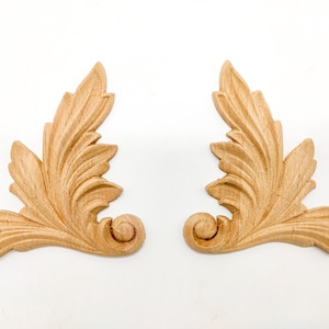 May include: Two ornate wooden leaf-shaped embellishments with a scroll design at the bottom. The embellishments are light brown and have a natural wood finish.
