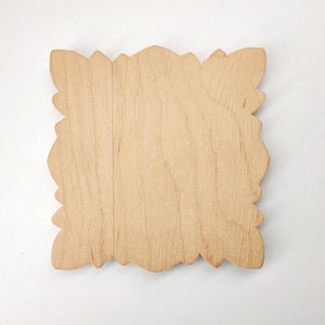 Puede incluir: Un letrero de madera en blanco con un borde decorativo y adornado. El letrero est&aacute; hecho de madera clara y tiene una superficie lisa y sin terminar.