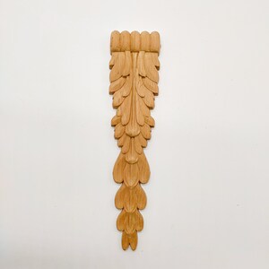 May include: A wooden carved ornament with a leaf design. The ornament is light brown and has a detailed, intricate design.