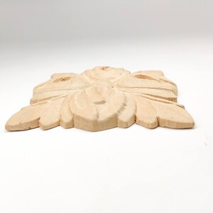 Puede incluir: Un adorno decorativo de madera marr&oacute;n claro con un dise&ntilde;o floral tallado. El adorno tiene forma de cuadrado con esquinas redondeadas.