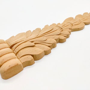 May include: A wooden carved leaf design with a detailed, intricate pattern. The design is light brown in color and has a smooth, polished finish.