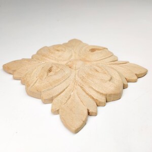 Puede incluir: Un adorno decorativo de madera marr&oacute;n claro con un dise&ntilde;o floral tallado. El dise&ntilde;o presenta cuatro hojas grandes y estilizadas con detalles intrincados.