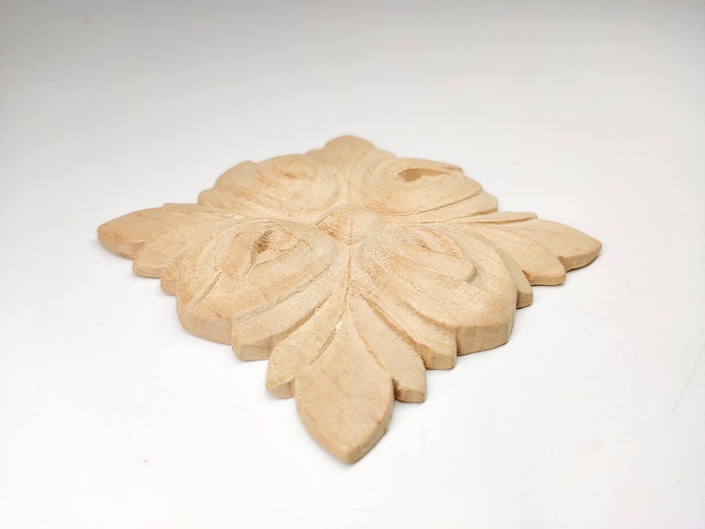 Puede incluir: Un adorno decorativo de madera marr&oacute;n claro con un dise&ntilde;o floral tallado. El dise&ntilde;o presenta cuatro hojas grandes y estilizadas con detalles intrincados.