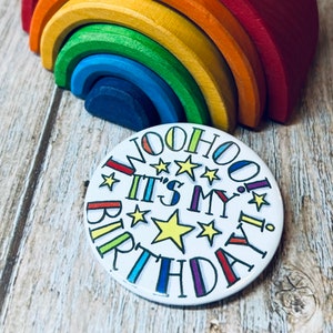 May include: A round button with a white background and colourful text that says "Woohoo! It's My Birthday!" with stars and a large yellow star in the centre.