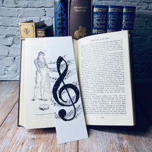 May include: A black and white bookmark with a treble clef design made of small circles, set against musical notation. The bookmark is in an open book with text, a classic literary theme.