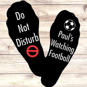 May include: Black socks with white text: "Do Not Disturb" with a red prohibition symbol and "Paul's Watching Football" with a football graphic. Set against a light wood grain background.