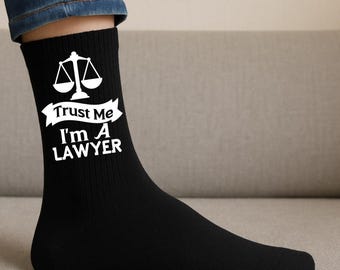May include: Black socks with the text "Trust Me I'm A LAWYER" in white, along with a white scale of justice graphic. The socks are ribbed and worn on a foot, with a portion of blue jeans visible.