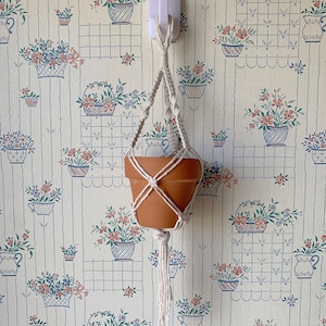 Puede incluir: Un colgador de plantas de macramé blanco con una maceta de terracota colgando de un gancho en una pared con un patrón de papel tapiz floral.