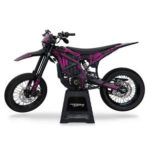 Può includere: Moto da cross elettrica nera e rosa con un design grafico rosa e nero. La moto è su un cavalletto nero con la scritta "Racing" stampata su di esso.
