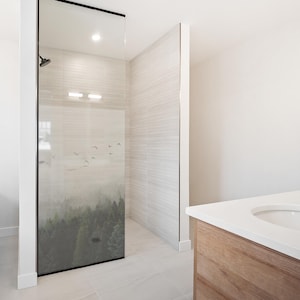 Op de afbeelding: Een moderne douchecabine met een glazen paneel met een boslandschap met vliegende vogels. De doucheruimte heeft lichtgekleurde tegelwanden en vloeren. Een houten wastafel met een wit aanrechtblad is zichtbaar in de hoek.