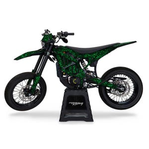 Op de afbeelding: Een zwarte en groene dirt bike met een groen en zwart camouflagepatroon. De motor staat op een zwarte standaard met het woord "Racing" erop.
