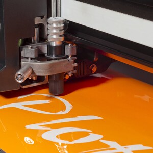 Puede incluir: Primer plano de una m&aacute;quina de corte negra y plateada con una perilla plateada y una cuchilla negra. La m&aacute;quina est&aacute; cortando un trozo de vinilo naranja con la palabra "Plot" escrita en blanco.