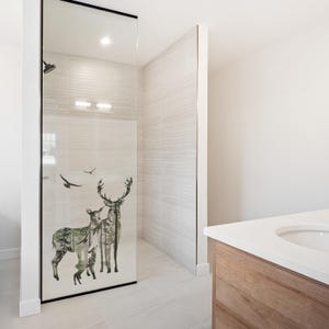 Könnte beinhalten: Eine rahmenlose Glas-Duschabtrennung mit einem Naturmotiv, das Hirsche und Vögel zeigt. Die Kunstwerke sind in Grün- und Grautönen gehalten und vor einem hellen Hintergrund platziert. Der Duschbereich hat weiße Fliesenwände und einen Holzwaschtisch.