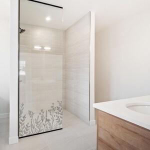 Peut inclure: Une porte de douche en verre sans cadre avec une bordure noire et un motif floral en bas. La zone de douche est carrelée de couleurs claires sur les murs et le sol. Un comptoir blanc avec un évier est visible.