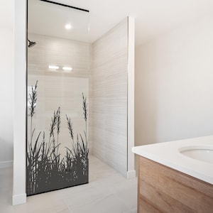 Può includere: Una porta della doccia in vetro trasparente con una silhouette nera di erba alta e canne. La porta della doccia è incorniciata in nero e fa parte di un moderno design del bagno. Il bagno ha pareti bianche e un mobile lavabo in legno.