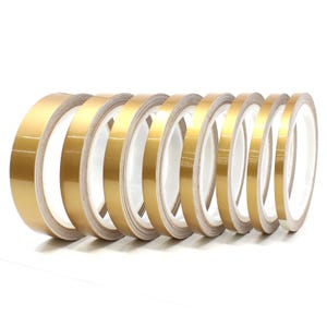 Op de afbeelding: Een verzameling gouden metallic tape rollen, elk met een glanzend, reflecterend oppervlak. De tape is opgerold op witte kernen, wat een contrast creëert. De rollen variëren enigszins in breedte.
