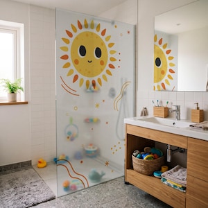 Op de afbeelding: Een douchewand van matglas met een grote, vrolijke gele zon met oranje stralen, kleine sterretjes en golvende lijnen. Het ontwerp is op het oppervlak gedrukt en belemmert gedeeltelijk het zicht op kleurrijk badspeelgoed in de douche.